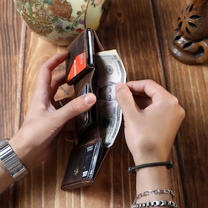 May include: A brown leather bi-fold wallet with multiple card slots and a cash compartment. The wallet is open, revealing a credit card and a US five-dollar bill. The wallet is held by two hands, showcasing its design and functionality.