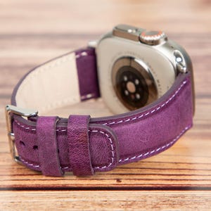 May include: A purple leather watch band with white stitching. The band is attached to a silver Apple Watch.