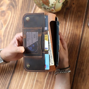 May include: A brown leather wallet with multiple card slots and a phone compartment. The wallet is open, revealing cards, cash, and a smartphone. The leather has a distressed finish, and the wallet is secured with snap closures.