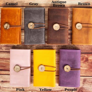 May include: Seven leather wallets in different colors: camel, gray, antique brown, brown, pink, yellow, and purple. Each wallet has a wooden button closure and a leather strap.
