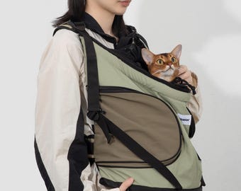 May include: A tan baseball cap with the word "COLOR" printed on the front. A cat carrier in olive green and brown with black straps. A cat with orange and brown fur is inside the carrier.