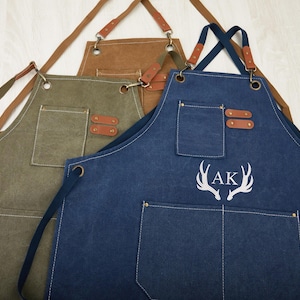 May include: Three canvas aprons with adjustable straps and pockets. One apron is green, one is brown, and one is blue with the initials "AK" embroidered on it.