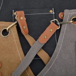 May include: A black, brown, and gray canvas apron with adjustable straps and metal hardware. The apron has two large pockets and a smaller pocket on the chest.