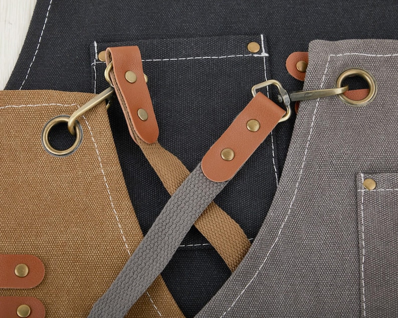 May include: A black, brown, and gray canvas apron with adjustable straps and metal hardware. The apron has two large pockets and a smaller pocket on the chest.