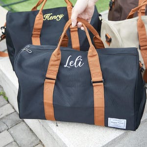 Peut inclure: Un sac de voyage noir avec des poign&eacute;es en tissu marron et le pr&eacute;nom Leli brod&eacute; en blanc sur le devant. Le sac pr&eacute;sente une fermeture &eacute;clair argent&eacute;e et une petite &eacute;tiquette rectangulaire blanche dans le coin avec le texte Happy Travel.