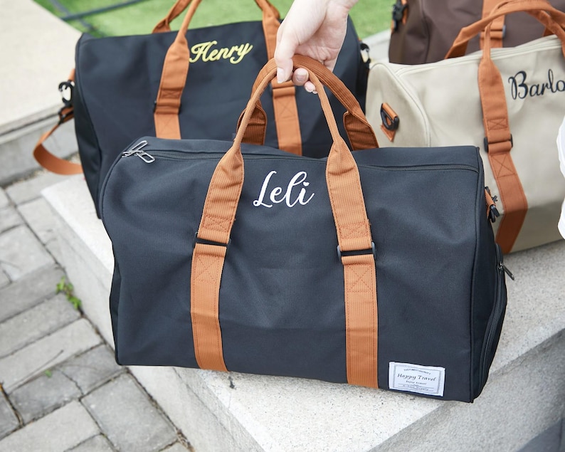 Peut inclure: Un sac de voyage noir avec des poign&eacute;es en tissu marron et le pr&eacute;nom Leli brod&eacute; en blanc sur le devant. Le sac pr&eacute;sente une fermeture &eacute;clair argent&eacute;e et une petite &eacute;tiquette rectangulaire blanche dans le coin avec le texte Happy Travel.