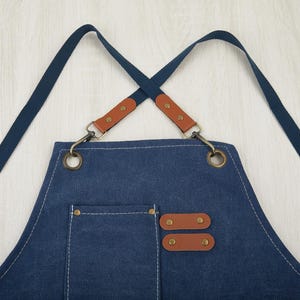 May include: A blue denim apron with brown leather straps and adjustable buckles. The apron has a large pocket on the front and two smaller pockets on the sides.