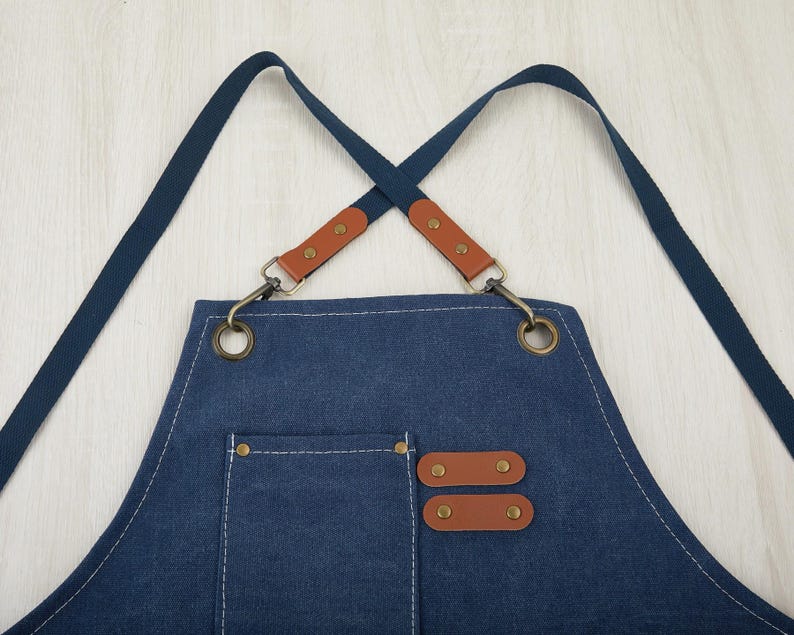 May include: A blue denim apron with brown leather straps and adjustable buckles. The apron has a large pocket on the front and two smaller pockets on the sides.