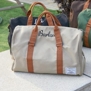 Peut inclure: Un sac de voyage beige avec des poign&eacute;es marron et une fermeture &eacute;clair. Le sac pr&eacute;sente le nom Barlow brod&eacute; en &eacute;criture noire sur le devant et un &eacute;cusson rectangulaire blanc avec le texte Happy Travel. D'autres sacs sont visibles en arri&egrave;re-plan.