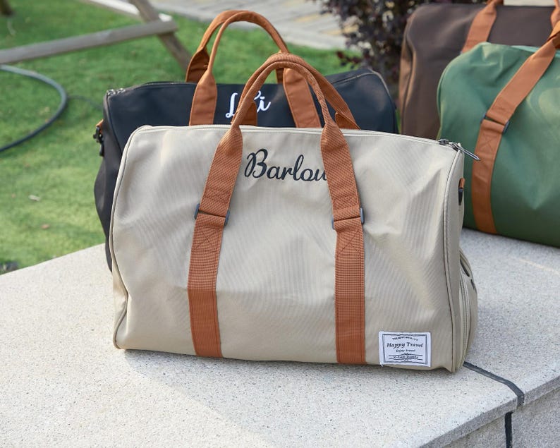 Peut inclure: Un sac de voyage beige avec des poign&eacute;es marron et une fermeture &eacute;clair. Le sac pr&eacute;sente le nom Barlow brod&eacute; en &eacute;criture noire sur le devant et un &eacute;cusson rectangulaire blanc avec le texte Happy Travel. D'autres sacs sont visibles en arri&egrave;re-plan.