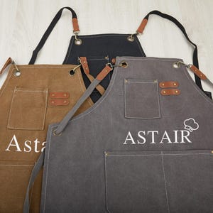 Könnte beinhalten: Drei Schürzen aus Canvas mit verstellbaren Trägern und Lederakzenten. Die Schürzen sind braun, grau und schwarz. Die graue Schürze hat das Wort "ASTAIR" auf der Vorderseite gestickt.