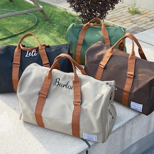 May include: Four duffel bags in black, olive green, brown, and beige, each with brown handles and straps. The bags have a rectangular shape and feature the names "Leli" and "Barla" embroidered in white. The bags are placed on a concrete surface.
