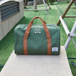 May include: Green duffel bag with brown handles and straps. The bag features a white embroidered design of crossed golf clubs and the letters "B" and "F". A small white label is on the lower right corner.