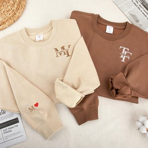 Könnte beinhalten: Zwei Sweatshirts, eines beige und eines braun, mit gestickten Initialen und einem kleinen Herzen auf dem beigen Sweatshirt. Das beige Sweatshirt hat die Initialen "M.Y." auf der Vorderseite gestickt und ein kleines rotes Herz auf dem Ärmel. Das braune Sweatshirt hat die Initialen "T.F." auf der Vorderseite gestickt.