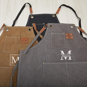 May include: Three canvas aprons with adjustable straps and leather accents. One apron is brown, one is grey, and one is black. The grey apron has the name "Martin" embroidered on it.