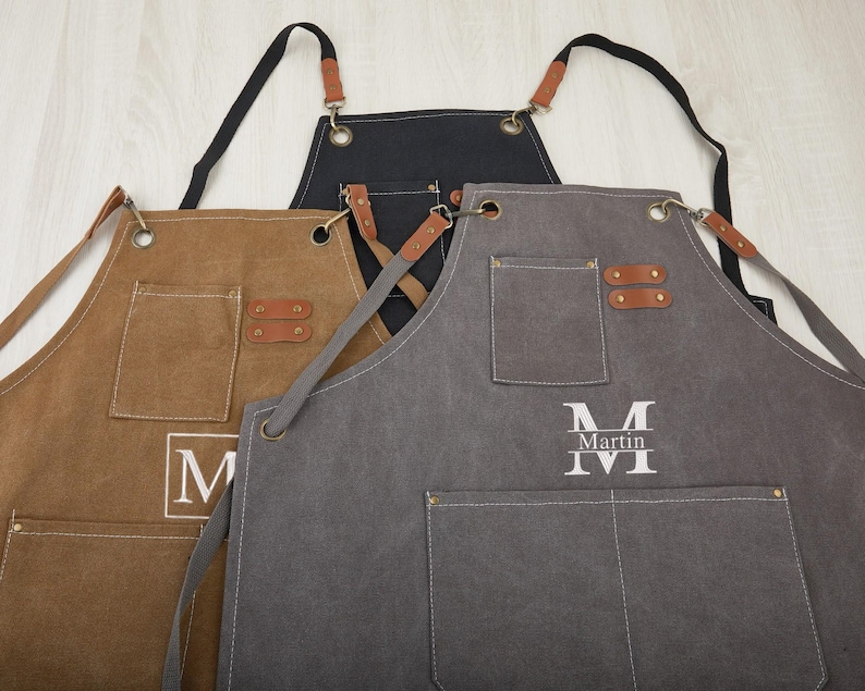 May include: Three canvas aprons with adjustable straps and leather accents. One apron is brown, one is gray, and one is black. The gray apron has the name "Martin" embroidered on it.