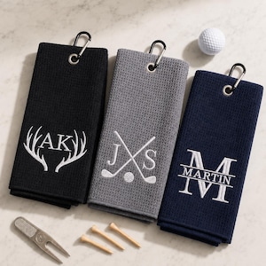May include: Three rectangular waffle-weave golf towels in black, grey, and navy blue, each featuring a metal carabiner clip. The black towel has white embroidered antlers and initials AK. The grey towel shows crossed golf clubs and initials JS. The navy towel displays the name MARTIN with initials M. A white golf ball, three wooden tees, and a metal divot tool are included.