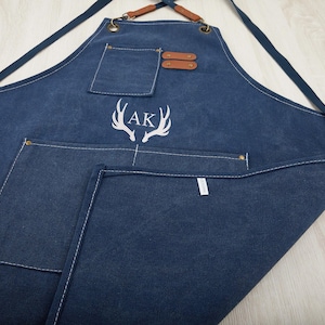 May include: A dark blue denim apron with a large pocket and a smaller pocket on the front. The apron has adjustable straps and a leather detail at the top. The apron is embroidered with the initials "AK" and a pair of antlers.