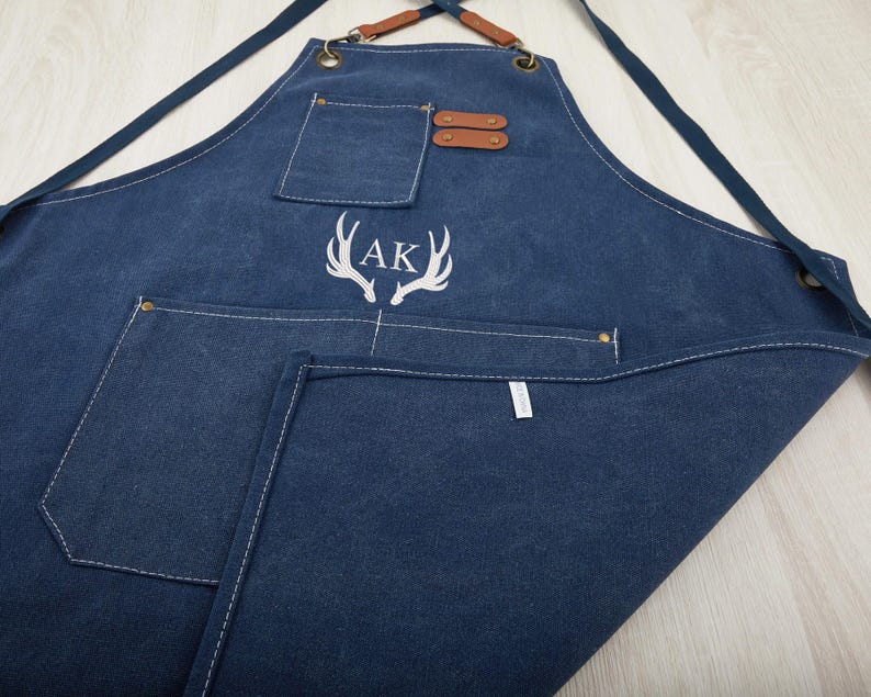 May include: A dark blue denim apron with a large pocket and a smaller pocket on the front. The apron has adjustable straps and a leather detail at the top. The apron is embroidered with the initials "AK" and a pair of antlers.
