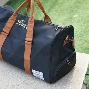Peut inclure: Un sac de sport noir avec des poign&eacute;es et des bordures en tissu beige. Le sac pr&eacute;sente le nom Henry brod&eacute; en lettres dor&eacute;es sur le c&ocirc;t&eacute;, une fermeture &eacute;clair sup&eacute;rieure, un compartiment lat&eacute;ral zipp&eacute; et une &eacute;tiquette blanche Happy Travel.