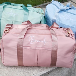 Peut inclure: Trois sacs de sport personnalisés de couleurs pastel : rose, vert menthe et bleu clair. Chaque sac porte un nom brodé en écriture blanche. Les sacs ont des poignées et des sangles assorties, et une quincaillerie dorée.