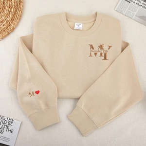 May include: A beige sweatshirt with a brown embroidered design that says "MY December 25th" and a red heart embroidered on the left sleeve.