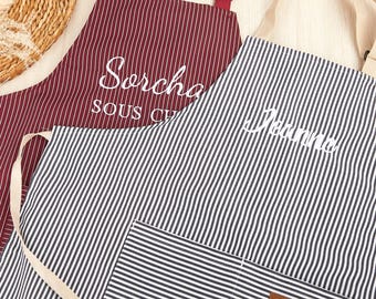 May include: Two striped aprons. One is burgundy with white pinstripes and the name "Sorcha" in cursive. The other is black and white striped with the name "Jeanne" in cursive. Both have pockets and adjustable straps.