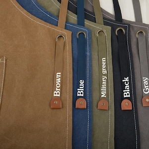 May include: Five canvas aprons with adjustable straps in brown, blue, olive green, black, and gray. Each apron has a white label with the color name printed on it.