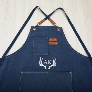 May include: A blue denim apron with adjustable straps and a large pocket. The apron has a white embroidered design of antlers with the initials "AK" in the center.