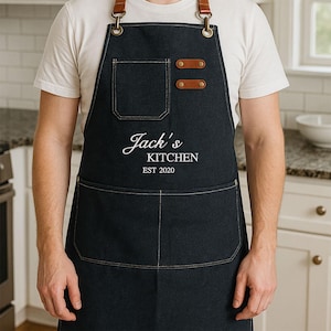 May include: A black denim apron with brown leather straps and accents. The apron has a large front pocket and a smaller chest pocket. The text "Jack's KITCHEN EST 2020" is printed in white script on the front.