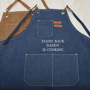 Puede incluir: Dos delantales, uno marrón y otro azul marino, con detalles de cuero y bolsillos. El delantal azul marino tiene el texto bordado "STAND BACK DADDY IS COOKING". Ambos delantales tienen correas ajustables.