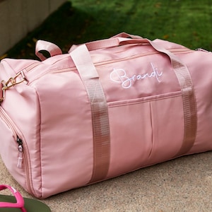 May include: A pink duffel bag with the name "Brandi" embroidered in white script. The bag has a zippered side pocket, top handles, and a shoulder strap. The bag is made of a smooth, durable material and is designed for travel or gym use.
