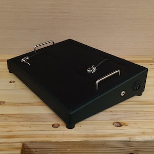 May include: A black rectangular effects pedalboard with two silver handles and a power input. The pedalboard has a textured surface and is designed to hold guitar effects pedals. The background is a wooden surface.