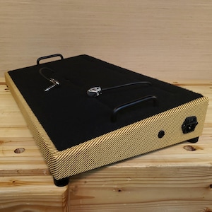 May include: A black pedalboard with a tweed-style frame and black handles. The top is covered in black material, and it has a power input and a jack input. The pedalboard is designed for use with musical instruments.