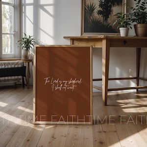 May include: A framed print with a warm terracotta background and the text "The Lord is my shepherd; I shall not want" in elegant white script. The print is displayed on a wooden floor, with a wooden table and plants in the background.