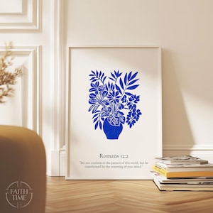 May include: A framed art print featuring a blue floral arrangement in a vase. The artwork includes the text "Romans 12:2" and a quote. The print is displayed on a wooden floor, leaning against a white wall.