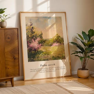 May include: A framed landscape art print with a beige border, leaning against a wall. The artwork features a colorful, impressionistic scene with pink and green foliage. Text at the bottom reads "Psalm 121:7-8". A wooden cabinet and potted plants are also visible.