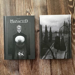May include: Two hardcover books with dark covers. The book on the left has the title "MANACLED" and a grayscale illustration of a person. The book on the right features a grayscale illustration of figures walking on a path.