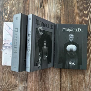 Op de afbeelding: Een verzameling grijze hardcover boeken met de titel "Manacled". De boekomslagen tonen illustraties van personages in donkere tinten. Eén boek is verpakt in papier met patronen en vastgebonden met touw.