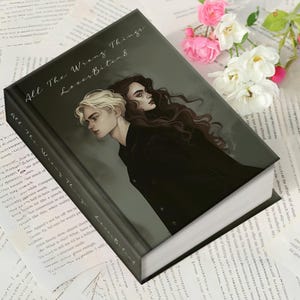 May include: A hardcover book titled "All The Wrong Things Loves Bitend" with a dark cover featuring two illustrated figures. The book is surrounded by printed pages and flowers, suggesting a literary theme.