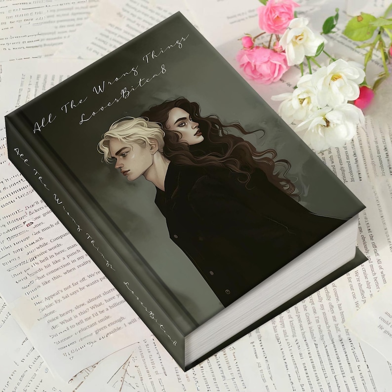 May include: A hardcover book titled "All The Wrong Things Loves Bitend" with a dark cover featuring two illustrated figures. The book is surrounded by printed pages and flowers, suggesting a literary theme.