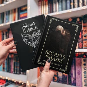 May include: Two black hardcover books with the title "Secrets & Masks" in gold lettering. The book on the right has a gold ornate border and a black and white illustration of two people embracing.