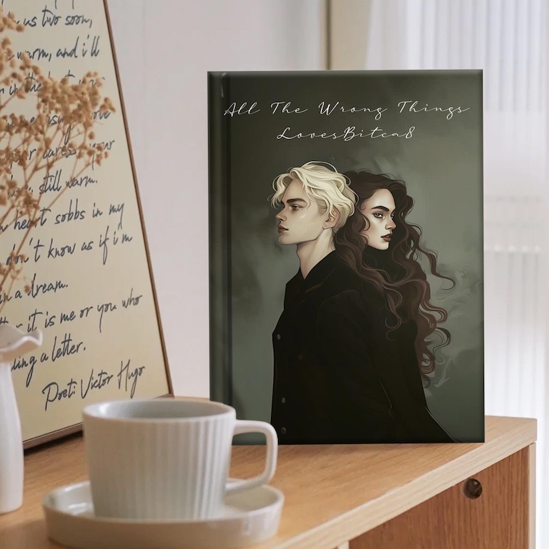 May include: A book cover with the title "All The Wrong Things LovesBitead" and an illustration of two people. One has blonde hair, the other has dark hair. The background is a muted green. A white teacup and saucer are in the foreground.