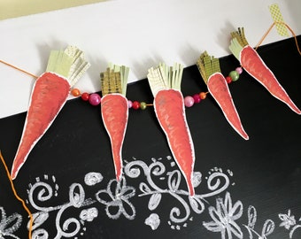 May include: A garland of five orange paper carrots with green tops strung together with a string of colorful beads. The garland is hanging on a black chalkboard with white chalk drawings of flowers and swirls.