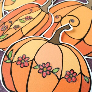May include: Three orange pumpkins with black outlines and glitter accents. Each pumpkin has a different design with small pink flowers and green leaves.