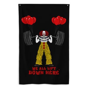 May include: A black rectangular wall banner featuring a clown with red hair, a ruffled collar, and yellow pants lifting a barbell with red balloons attached to each side. The text WE ALL LIFT DOWN HERE is printed in red at the bottom of the banner.