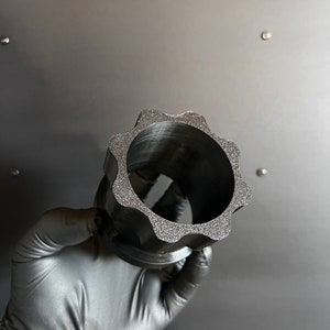 May include: A black plastic gear-shaped object with a textured surface. The object has a large hole in the center.
