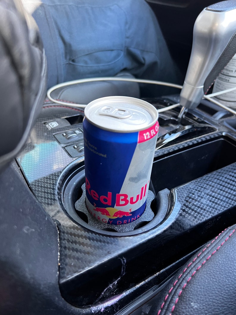 May include: A red and blue Red Bull energy drink can in a black cup holder in a car. The can has the text "Red Bull" and "Energy Drink" on it.