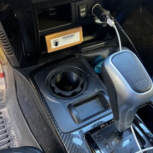 May include: A black car interior with a black and silver gear shifter, a black cup holder, and a small brown box with the text "X22.02.007" on it. The box is in a small compartment above the cup holder. There is a white USB cable and a white AUX cable plugged into the car's dashboard.