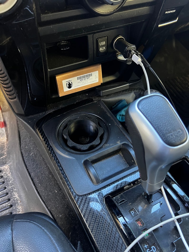 May include: A black car interior with a black and silver gear shifter, a black cup holder, and a small brown box with the text "X22.02.007" on it. The box is in a small compartment above the cup holder. There is a white USB cable and a white AUX cable plugged into the car's dashboard.